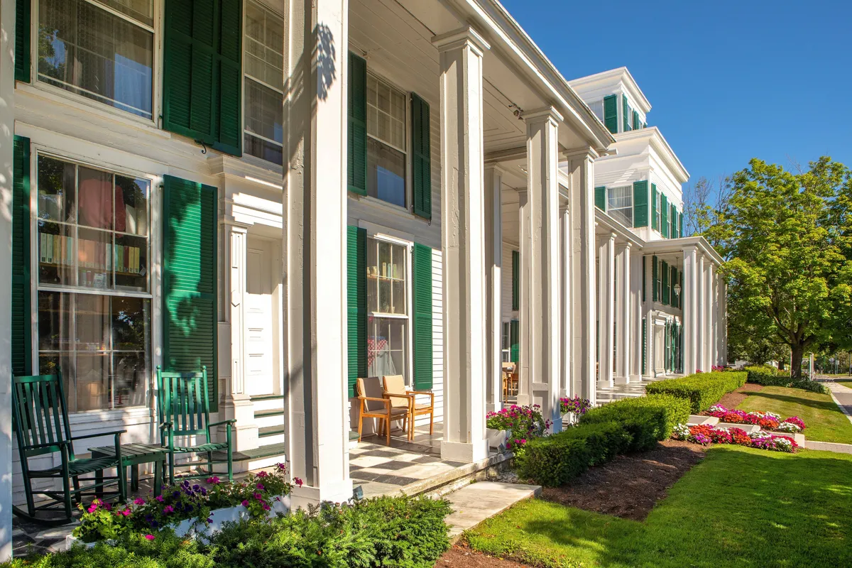 A welcoming New England-style porch with classic green shutters, white columns, and flower beds. Two traditional rocking chairs sit side by side, inviting guests to relax and enjoy the vibrant porch scene. The soft morning light enhances the inviting atmosphere, and the porch overlooks lush landscape, suggesting both tranquility and connection to the outdoors.