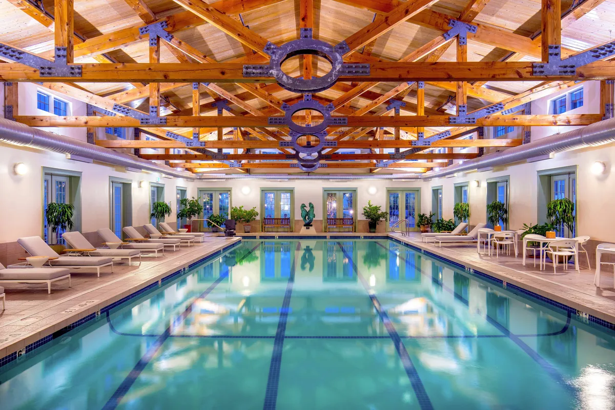 A serene indoor swimming pool accented by wooden beams, lounge chairs perfect for relaxing, tasteful sculptures, and lush potted plants. The inviting space offers a luxurious atmosphere ideal for unwinding and enjoying moments of calm.