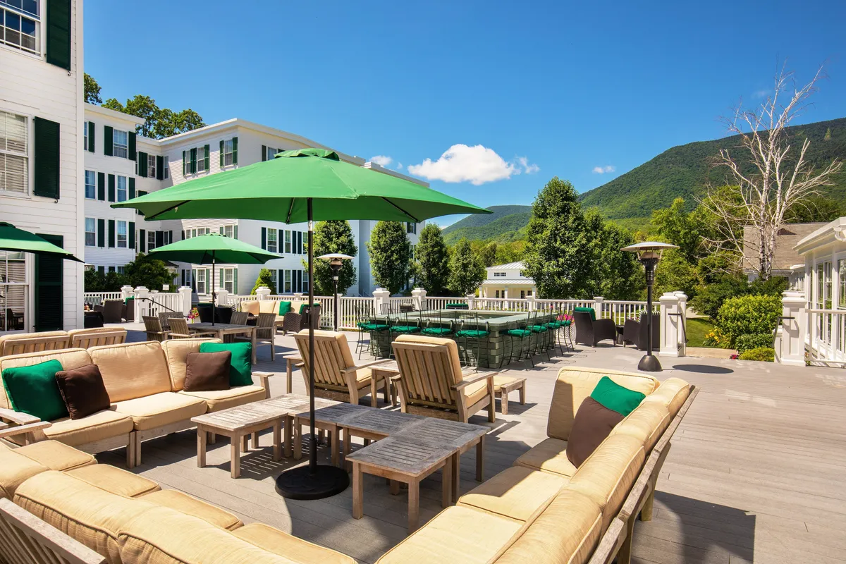 A cheerful outdoor terrace with comfortable sofas, green umbrellas, and dining tables, all oriented toward breathtaking mountain views. The space is inviting for social gatherings, relaxed afternoons, or enjoying a meal outdoors as the sun sets.