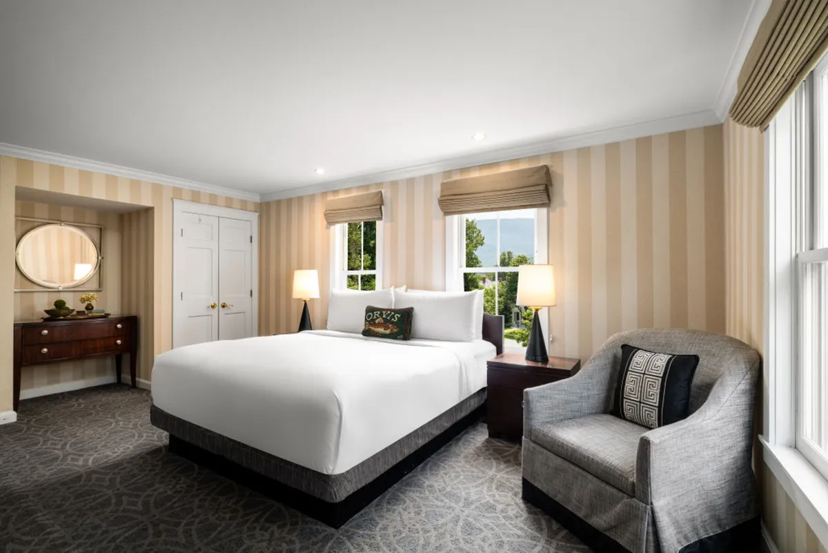 The image showcases a serene, sunlit bedroom featuring a plush bed, a cozy armchair by the window, and inviting natural light streaming in. The decor evokes comfort and classic New England charm, suggesting a peaceful retreat perfect for relaxation and unwinding. The overall atmosphere is calm and inviting, with soft textures and thoughtful details that highlight the property's luxury and warmth.