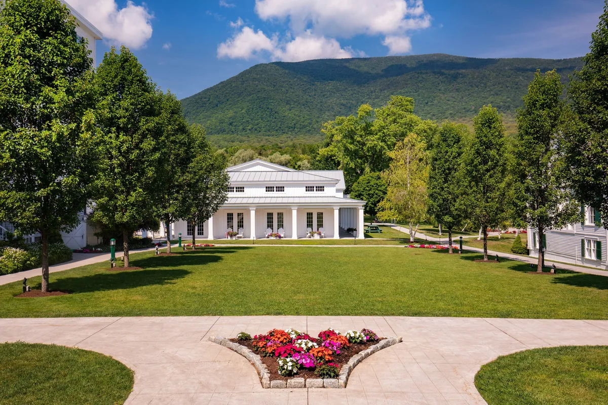A tranquil exterior view of a manicured lawn and vibrant flower beds, framed by the elegant architecture of a historic building. The scene extends toward a scenic mountain backdrop, with outdoor gathering spaces suggesting relaxation and celebration in nature.
