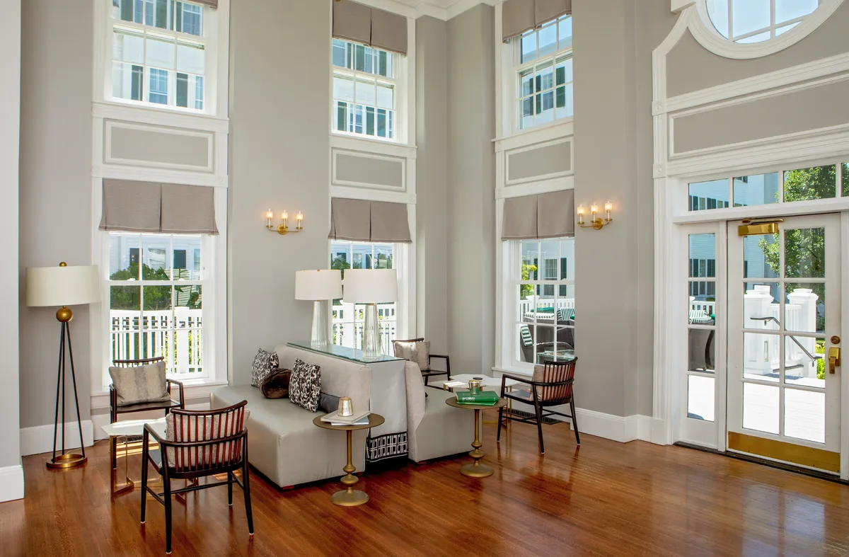 An elegant interior with large windows bathing the space in natural light, a stylish seating area, and a prominent entrance door. The scene suggests both a welcoming entry and a comfortable spot to pause and enjoy a drink or conversation.