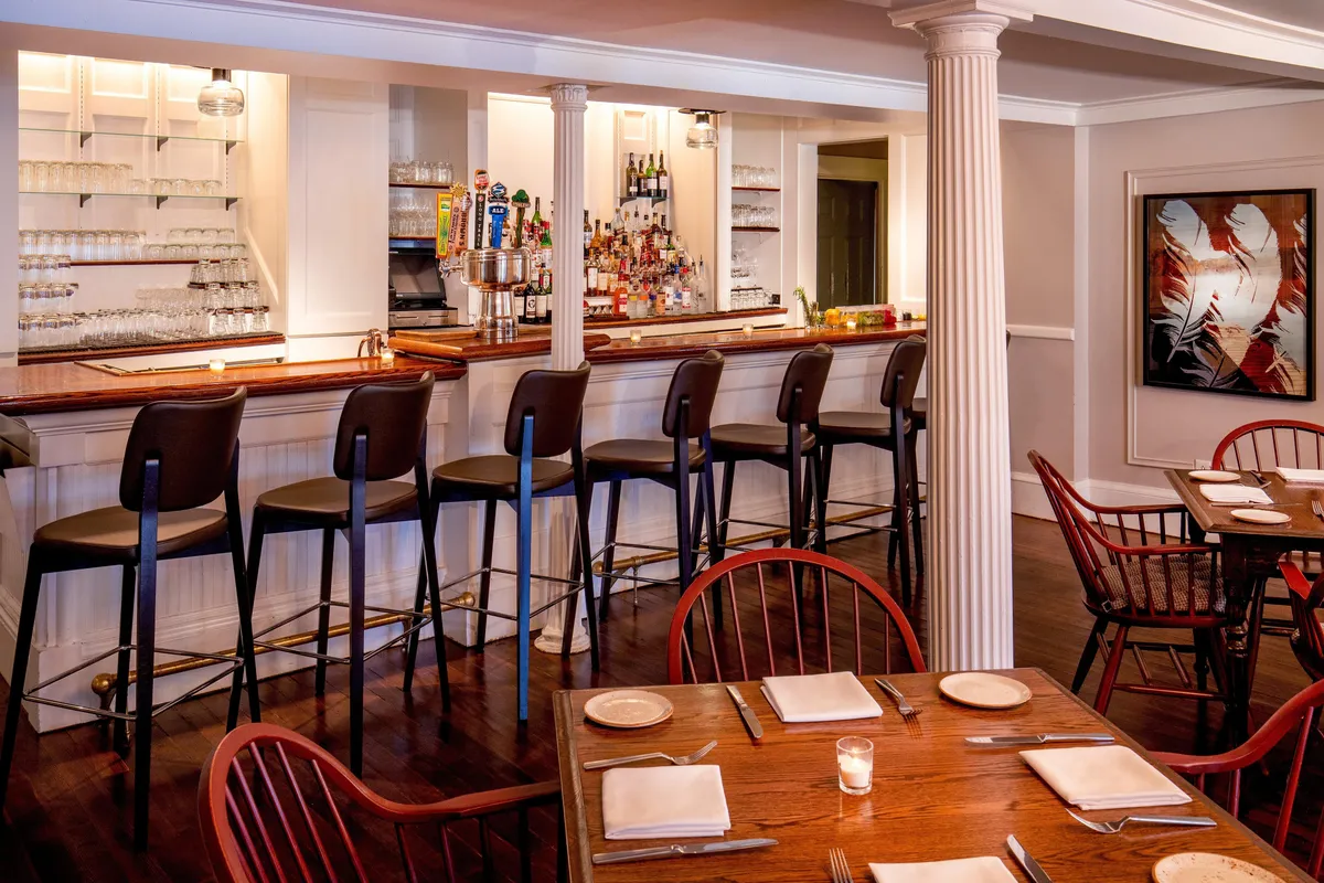 This image showcases a welcoming bar counter with comfortable stools, nearby dining tables, and vibrant artwork on the wall. The atmosphere is relaxed, with guests socializing and enjoying drinks or small plates.