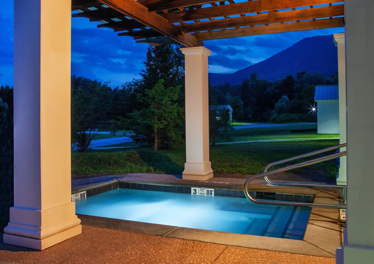 An illuminated pool at night beneath a stylish pergola, backed by the silhouette of mountains under a starlit sky. The scene evokes serenity and luxury, ideal for nighttime relaxation or romantic stargazing by the water.