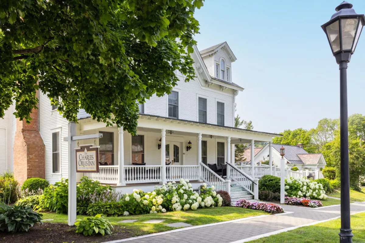 A broad front porch with white pillars overlooks colorful flower beds and features the sign 'CHARLES OVIS INN'. The porch is set against a backdrop of lush gardens and hints at the surrounding Vermont scenery. The scene captures the porch as an inviting bridge between the suite and the outdoors, with morning light and abundant flowers enhancing the sense of peace and welcome.