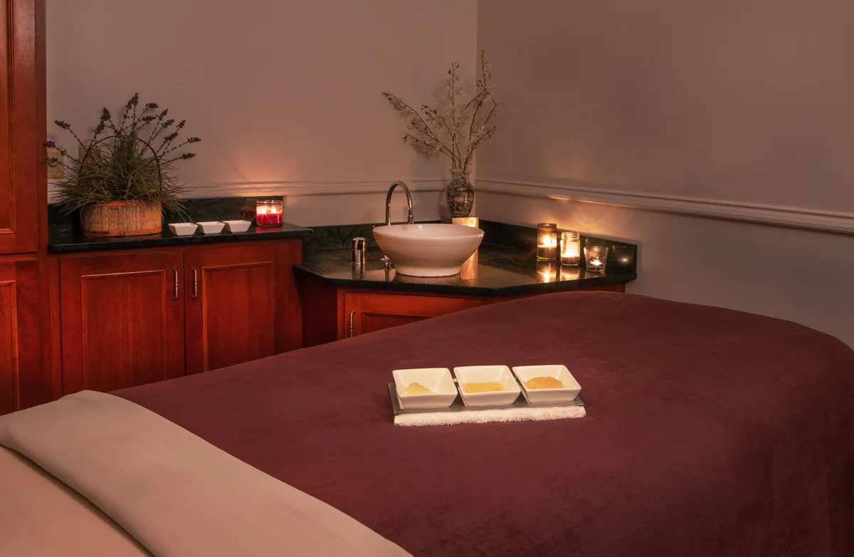 A tranquil interior spa space with a massage table, ceramic bowls, a vanity sink, and softly glowing candles, all set in a calm, inviting atmosphere ideal for self-care and wellness.